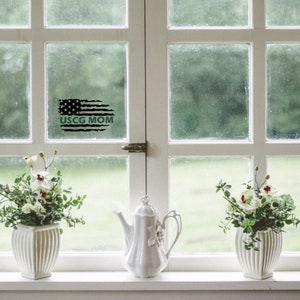 Op de afbeelding: Een raam met een zwart-witte Amerikaanse vlag sticker met de tekst "USCG MOM" in groen. Er staan twee witte keramische vazen met witte bloemen op een vensterbank.