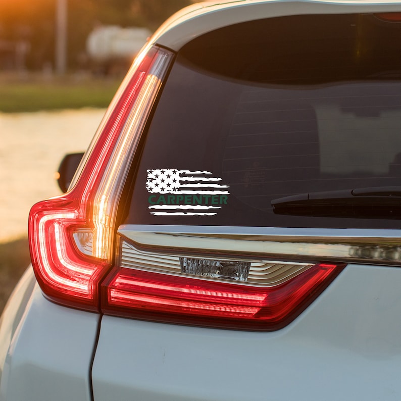 Puede incluir: Una calcoman&iacute;a de bandera estadounidense blanca desgastada con la palabra "CARPENTER" en texto verde en un coche plateado.