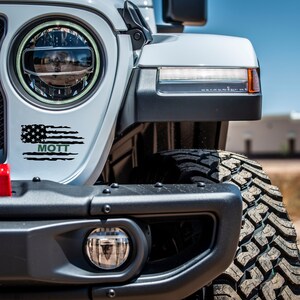 Puede incluir: Un Jeep Wrangler blanco con una calcoman&iacute;a de bandera estadounidense negra y verde que dice "MOTT". El Jeep tiene un parachoques delantero negro y una llanta negra.