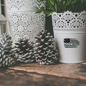 Op de afbeelding: Een witte keramische plantenpot met een kantontwerp, met een zwart-witte Amerikaanse vlag met de tekst "USCG MOM" in groen. De pot staat op een houten oppervlak met drie dennenappels.
