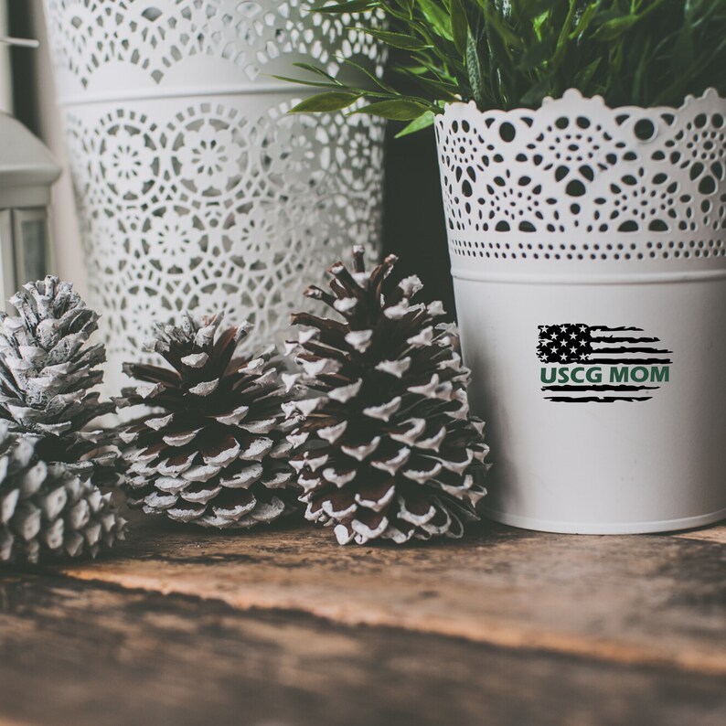 Op de afbeelding: Een witte keramische plantenpot met een kantontwerp, met een zwart-witte Amerikaanse vlag met de tekst "USCG MOM" in groen. De pot staat op een houten oppervlak met drie dennenappels.