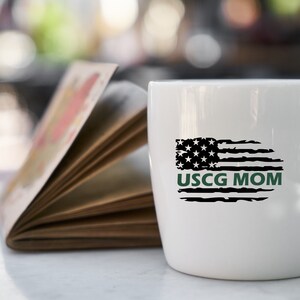 Op de afbeelding: Witte keramische mok met een verweerd Amerikaans vlagontwerp en de tekst "USCG MOM" in groene letters.