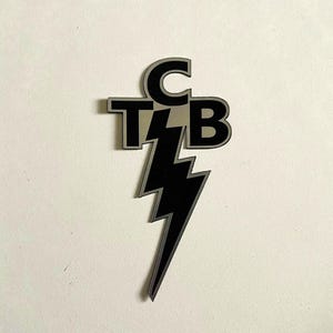 May include: A silver and black emblem featuring the letters "TCB" stacked with a lightning bolt design. The letters are in a bold, sans-serif font, and the lightning bolt is black with silver trim. The emblem is likely a decorative accessory.