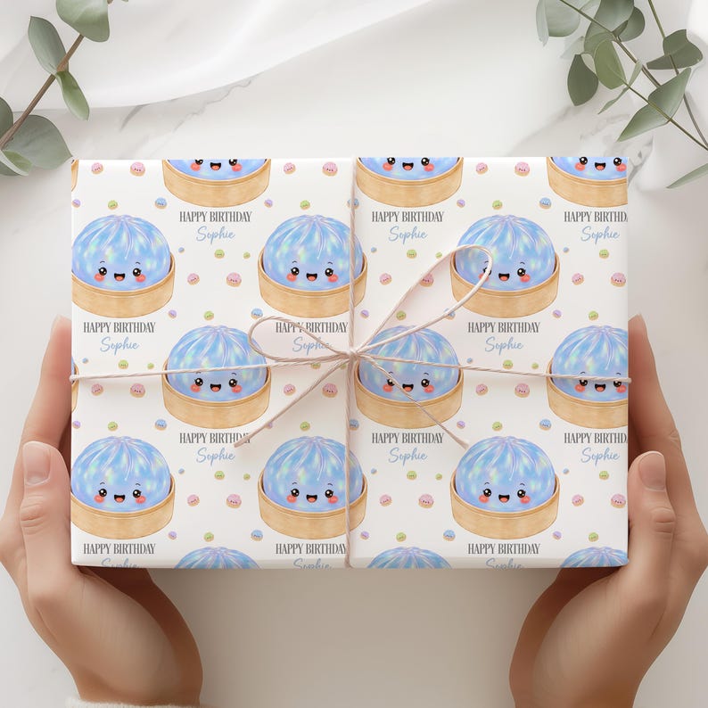 Op de afbeelding: Cadeaupapier met een herhaald patroon van lachende blauwe dumplings in bamboe stoommandjes, met de tekst "Happy Birthday Sophie" en kleurrijke confetti. Het cadeau is vastgebonden met een lichtbruin lint.