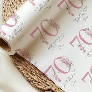 Op de afbeelding: Een rol wit cadeaupapier met een herhalend patroon voor een 70e verjaardag. Het ontwerp toont het getal 70 in een zachte roze kleur, versierd met bloemstukken. De tekst "Wishing you a very happy birthday" en de naam "Shona" zijn ook gedrukt.