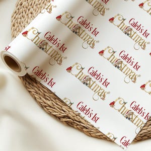 May include: White wrapping paper roll with a repeating pattern of "Caleb's 1st Christmas" in gold script and red text. The design includes teddy bears wearing Santa hats and snowflake accents.