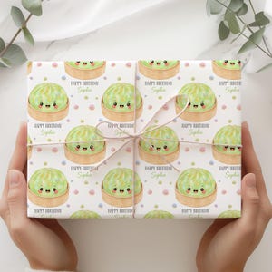 Op de afbeelding: Cadeaupapier met een herhalend patroon van cartoon dumplings in bamboe stoommandjes, met de tekst "HAPPY BIRTHDAY Sophie" op een witte achtergrond. Het cadeau is vastgebonden met een lichtgekleurd lint.