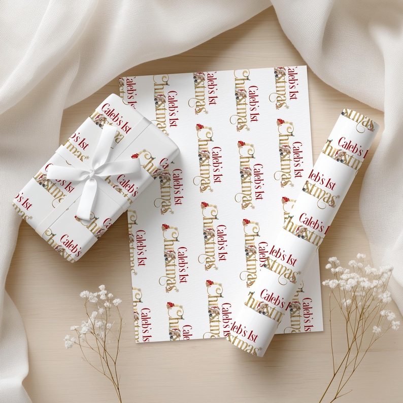 May include: White wrapping paper with a repeating pattern of the text "Caleb's 1st Christmas" in gold and red. A wrapped gift with a white ribbon, a sheet of paper, and a roll of paper are visible.