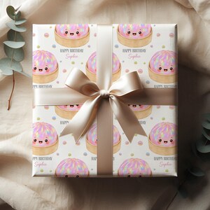 Op de afbeelding: Cadeaupapier met een herhaald patroon van cartoon bao buns in bamboe stoommandjes op wit papier. De tekst "HAPPY BIRTHDAY Sophie" is op de verpakking gedrukt. Een satijnen lint en strik maken het geheel af.