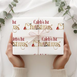 May include: A wrapped gift with the text "Caleb's 1st Christmas" in red and gold script. The wrapping paper features a repeating pattern of teddy bears and Christmas accents. The gift is tied with a light brown ribbon.