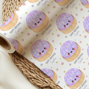 Op de afbeelding: Wit cadeaupapier met een herhaald patroon van lachende paarse bao-broodjes in bamboe stoommandjes. De tekst "HAPPY BIRTHDAY Sophie" staat op het papier. Pastelkleurige confetti stippen zijn verspreid.