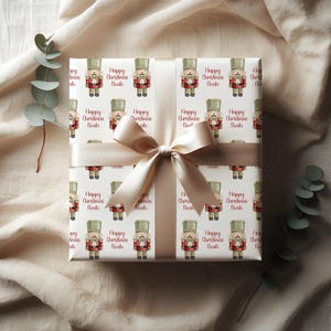 Op de afbeelding: Een cadeau verpakt in wit papier met een herhaald patroon van speelgoed notenkrakers in rood, groen en bruin. De woorden "Happy Christmas Noah" staan op het papier. Een crèmekleurig lint is in een strik geknoopt.