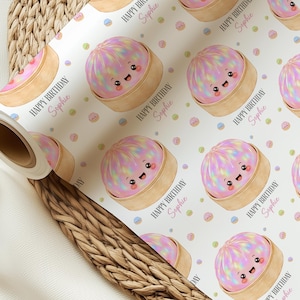 Op de afbeelding: Wit cadeaupapier met een herhaald patroon van lachende, regenboogkleurige bao-broodjes in bamboe stoommandjes. De tekst "HAPPY BIRTHDAY Sophie" is in roze gedrukt. Pastelkleurige confetti stippen maken het ontwerp compleet.