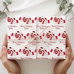 Pode incluir: Papel de embrulho branco com tema natalino com um padrão repetido de balões vermelhos com o número 67, chapéus de Papai Noel e o texto "Merry Christmas Dylan". O papel é amarrado com uma fita creme.