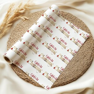 May include: A roll of white wrapping paper with a repeating pattern of the text "Caleb's 1st Christmas" in burgundy and gold. The design includes a Santa hat and festive illustrations. The paper is displayed on a woven mat.