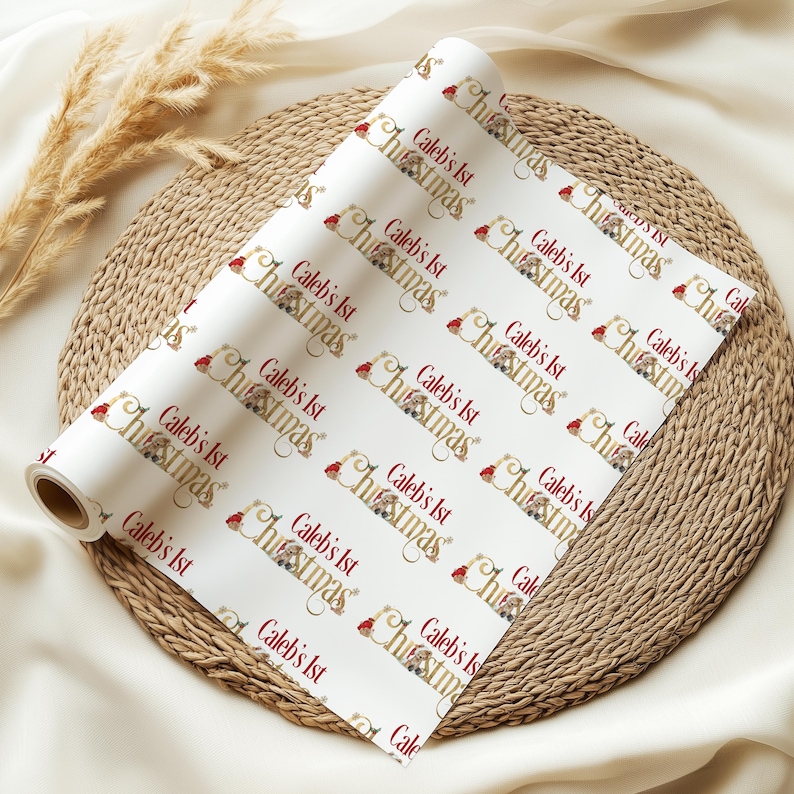 May include: A roll of white wrapping paper with a repeating pattern of the text "Caleb's 1st Christmas" in burgundy and gold. The design includes a Santa hat and festive illustrations. The paper is displayed on a woven mat.