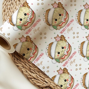 May include: A roll of white wrapping paper featuring a repeating pattern of cartoon gingerbread characters wearing Santa hats, accented with red and green sprinkles, and the text Merry Christmas Alicja printed in a red and brown script font.
