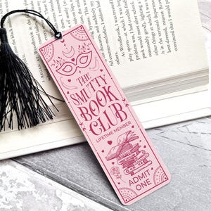Puede incluir: Un marcap&aacute;ginas rosa con el texto "THE SMUTTY BOOK CLUB" e ilustraciones decorativas. El marcap&aacute;ginas tiene una borla negra. El dise&ntilde;o incluye una m&aacute;scara, libros y las palabras "ADMIT ONE".