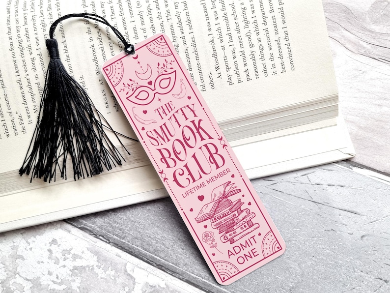 Puede incluir: Un marcap&aacute;ginas rosa con el texto "THE SMUTTY BOOK CLUB" e ilustraciones decorativas. El marcap&aacute;ginas tiene una borla negra. El dise&ntilde;o incluye una m&aacute;scara, libros y las palabras "ADMIT ONE".