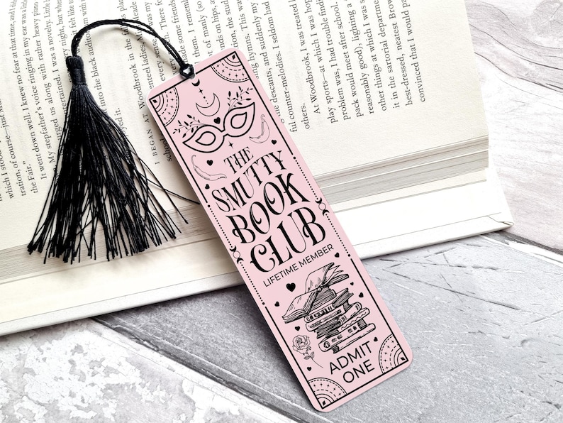 Puede incluir: Un marcap&aacute;ginas rosa con texto negro y una borla negra. El marcap&aacute;ginas presenta el texto "THE SMUTTY BOOK CLUB" e ilustraciones decorativas de una m&aacute;scara, libros y una rosa. Las palabras "ADMIT ONE" tambi&eacute;n est&aacute;n impresas.