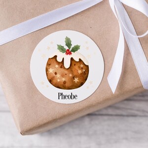 Pode incluir: Um autocolante redondo com uma ilustração em aquarela de um pudim de Natal, decorado com glacê branco, azevinho e estrelas. O nome "Phoebe" está impresso abaixo. O autocolante está num presente embrulhado com uma fita branca.
