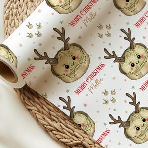 Può includere: Un rotolo di carta da regalo bianca con un motivo ripetuto di una tortina di frutta a cartone animato con corna di renna, guance rosate e luci natalizie. Il testo "MERRY CHRISTMAS Mollie" è stampato in rosso e oro tra piccoli fiocchi.
