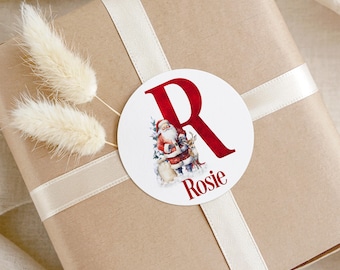 Könnte beinhalten: Ein runder weißer Aufkleber mit einem großen roten Buchstaben R, einer Aquarellillustration vom Weihnachtsmann mit einem Rentier und dem Namen Rosie in roter Schrift. Der Aufkleber klebt auf einem braunen Geschenkpaket mit cremefarbenem Band.