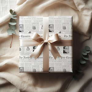 Pu&ograve; includere: Un regalo avvolto in carta da giornale con immagini di Babbo Natale e il testo "The North Pole Chronicle". &Egrave; legato con un nastro e un fiocco di raso color crema, su uno sfondo di tessuto neutro.