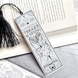 Op de afbeelding: Een witte bladwijzer met een zwart-witte illustratie van twee handen die in elkaar grijpen. De bladwijzer heeft de tekst "You had me at Trigger Warning" en "good girl" erop.
