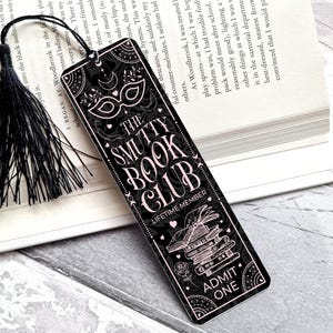 Puede incluir: Marcap&aacute;ginas negro con el texto "THE SMUTTY BOOK CLUB" en una fuente decorativa. El marcap&aacute;ginas tiene una borla negra y un dise&ntilde;o con una m&aacute;scara, libros y las palabras "ADMIT ONE".