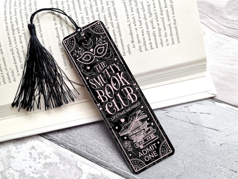 Puede incluir: Marcap&aacute;ginas negro con el texto "THE SMUTTY BOOK CLUB" en una fuente decorativa. El marcap&aacute;ginas tiene una borla negra y un dise&ntilde;o con una m&aacute;scara, libros y las palabras "ADMIT ONE".