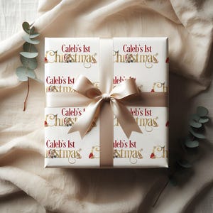 May include: A wrapped gift with white paper featuring the text "Caleb's 1st Christmas" in gold and red. The gift is tied with a cream-colored ribbon and bow. Eucalyptus sprigs are in the background.