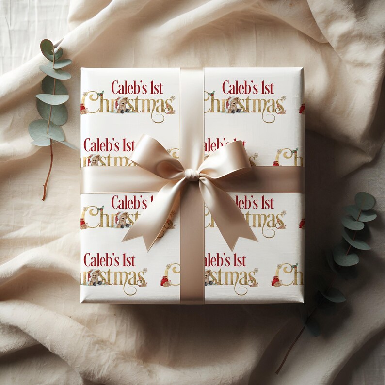 May include: A wrapped gift with white paper featuring the text "Caleb's 1st Christmas" in gold and red. The gift is tied with a cream-colored ribbon and bow. Eucalyptus sprigs are in the background.