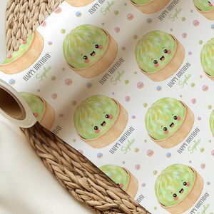 Op de afbeelding: Wit cadeaupapier met een herhalend patroon van cartoon dim sum in bamboe stoommandjes. De dim sum zijn lichtgroen met lachende gezichten. De tekst "HAPPY BIRTHDAY Sophie" wordt herhaald.