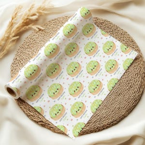 Op de afbeelding: Een rol wit cadeaupapier met een herhaald patroon van cartoon groene cakes met lachende gezichten. De woorden "Happy Birthday Sophie" staan op het papier gedrukt, samen met kleurrijke stippen.