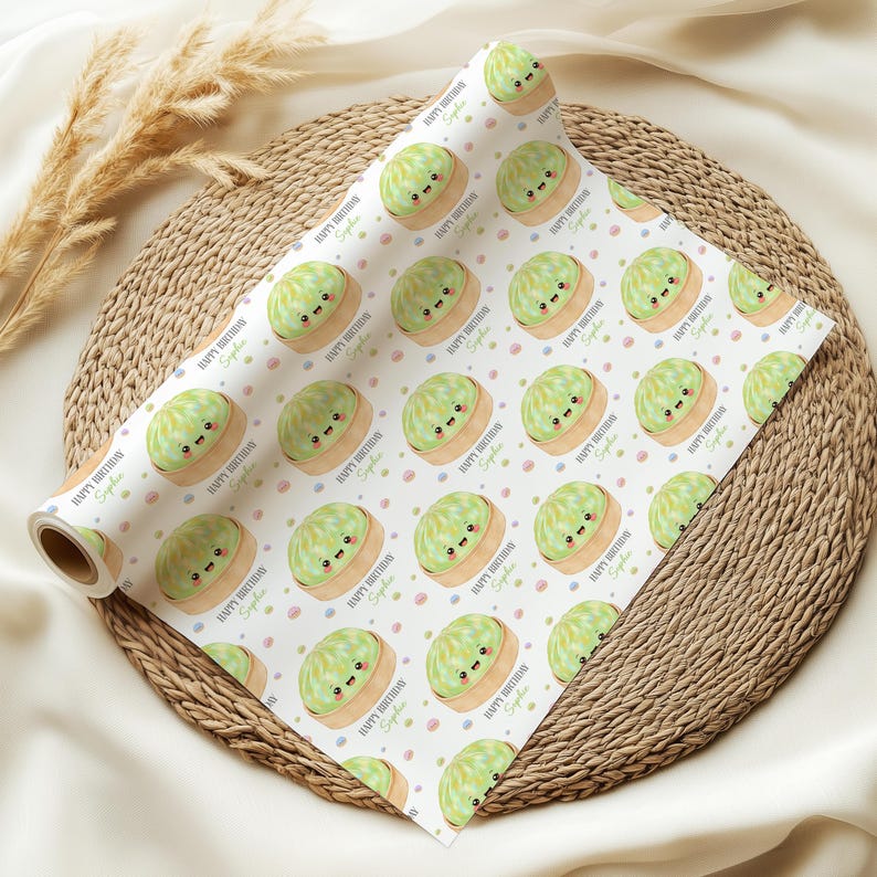Op de afbeelding: Een rol wit cadeaupapier met een herhaald patroon van cartoon groene cakes met lachende gezichten. De woorden "Happy Birthday Sophie" staan op het papier gedrukt, samen met kleurrijke stippen.