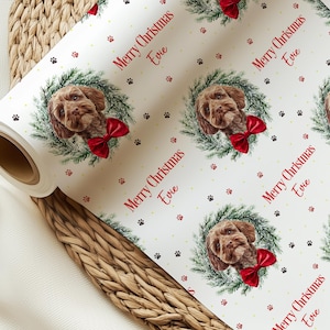 Pode incluir: Um rolo de papel de embrulho branco com um padrão repetitivo de um cão castanho com um laço vermelho, rodeado por uma coroa de pinheiro verde. O texto Merry Christmas Evie está impresso a vermelho, com pequenos ícones de pegadas.