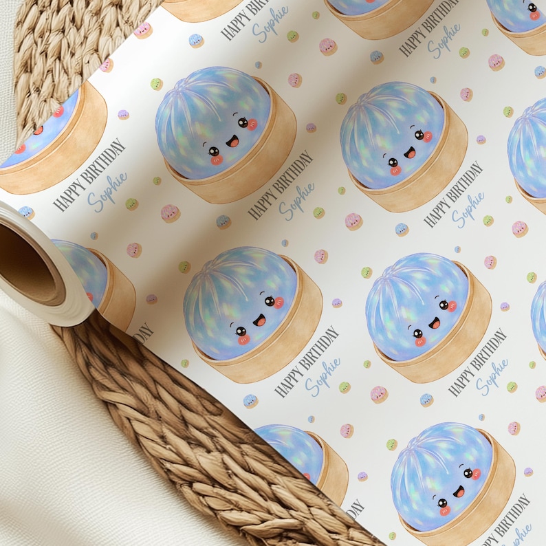 Op de afbeelding: Wit cadeaupapier met een herhaald patroon van lachende blauwe dumplings in bamboe stoommandjes. De woorden "HAPPY BIRTHDAY Sophie" zijn gedrukt in een scriptlettertype. Pastelkleurige confetti stippen zijn verspreid over de achtergrond.