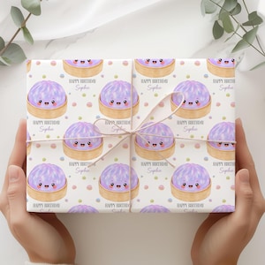 Op de afbeelding: Cadeaupapier met een herhaald patroon van lachende paarse dumplings in bamboe stoommandjes, met de tekst "Happy Birthday Sophie" op een witte achtergrond. Het cadeau is vastgebonden met touw.