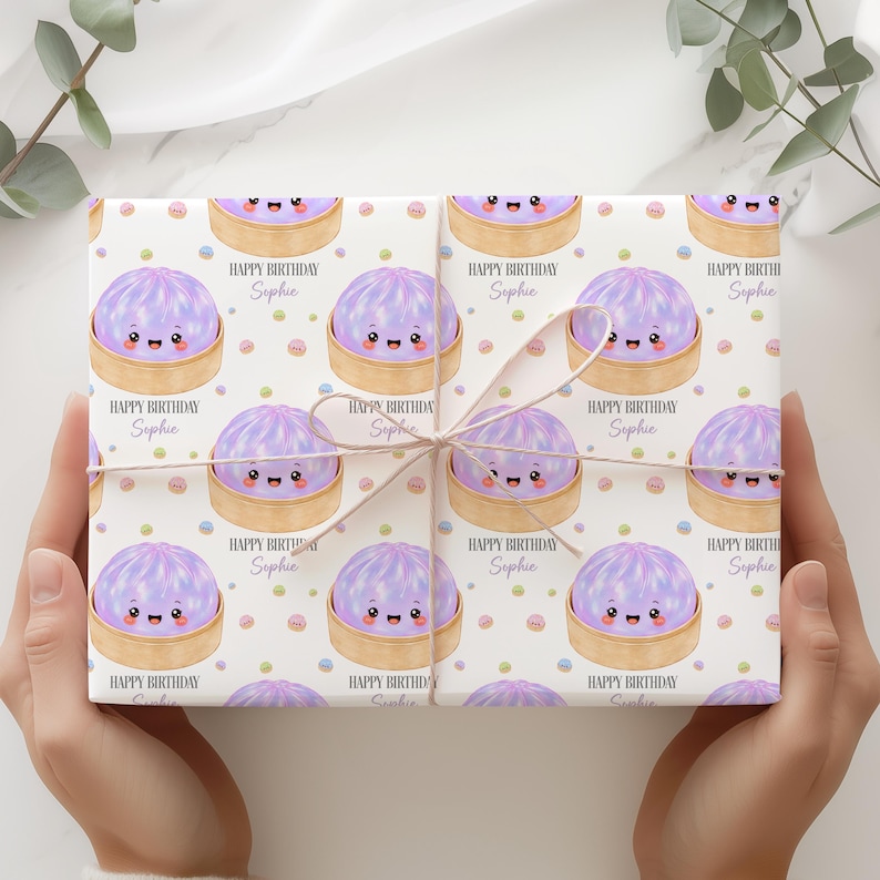 Op de afbeelding: Cadeaupapier met een herhaald patroon van lachende paarse dumplings in bamboe stoommandjes, met de tekst "Happy Birthday Sophie" op een witte achtergrond. Het cadeau is vastgebonden met touw.