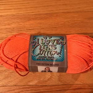 May include: A skein of bright orange yarn with a paper label that reads I LOVE THIS COTTON! in holographic lettering. The label features a blue polka dot background, the text SUPER SOFT, and a small photograph of a person wearing a garment.