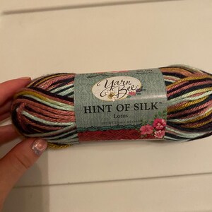 May include: A skein of Yarn Bee Hint of Silk yarn in the color Lotus. The yarn is a multi-colored blend of pink, green, yellow, black, and white.
