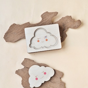 May include: Two wooden cloud shaped puzzle pieces. One piece is a cutout of a cloud with a smiling face and closed eyes. The other piece is a solid cloud with a smiling face and closed eyes. Both clouds have pink cheeks and one has a blue eye.