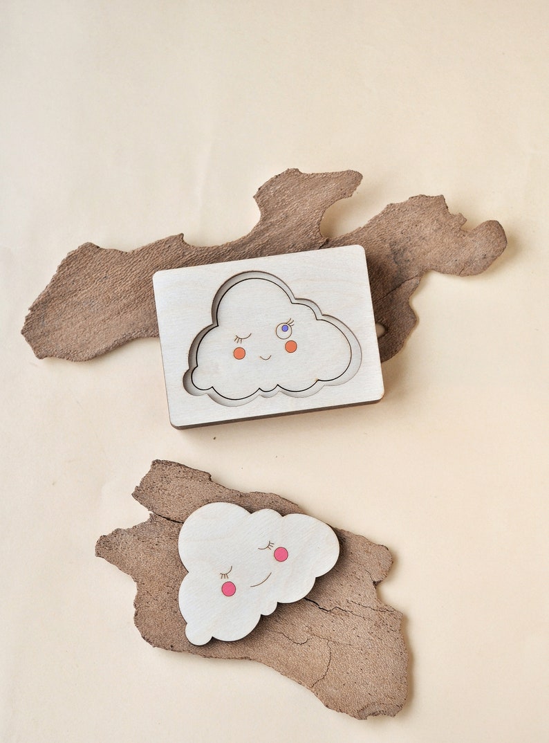 May include: Two wooden cloud shaped puzzle pieces. One piece is a cutout of a cloud with a smiling face and closed eyes. The other piece is a solid cloud with a smiling face and closed eyes. Both clouds have pink cheeks and one has a blue eye.