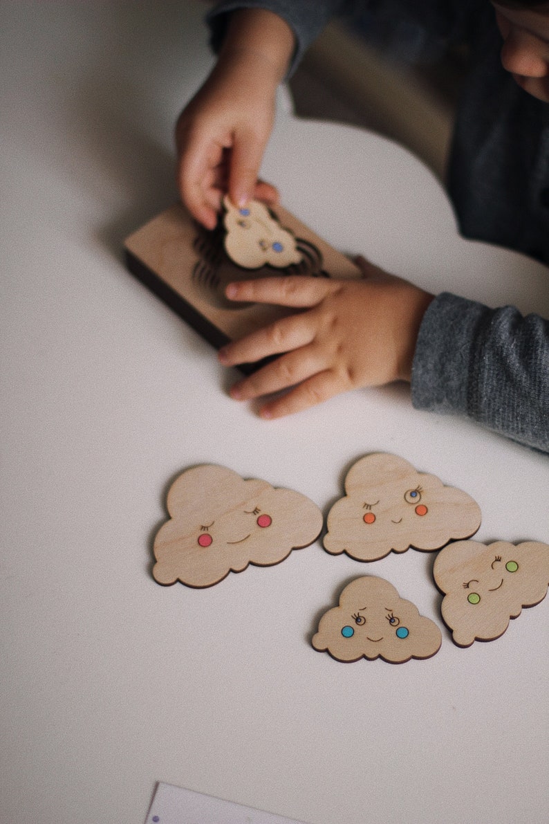 May include: A wooden puzzle with five cloud-shaped pieces, each with a different facial expression. The clouds have painted eyes, mouths, and cheeks in various colors. The puzzle is designed for children to learn about emotions.
