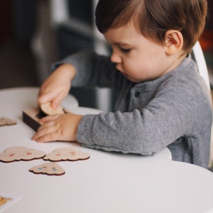 May include: A young child sits at a white, cloud-shaped table, playing with a wooden puzzle featuring colorful clouds. The puzzle pieces are arranged on the table, and the child is holding one piece in their hand.