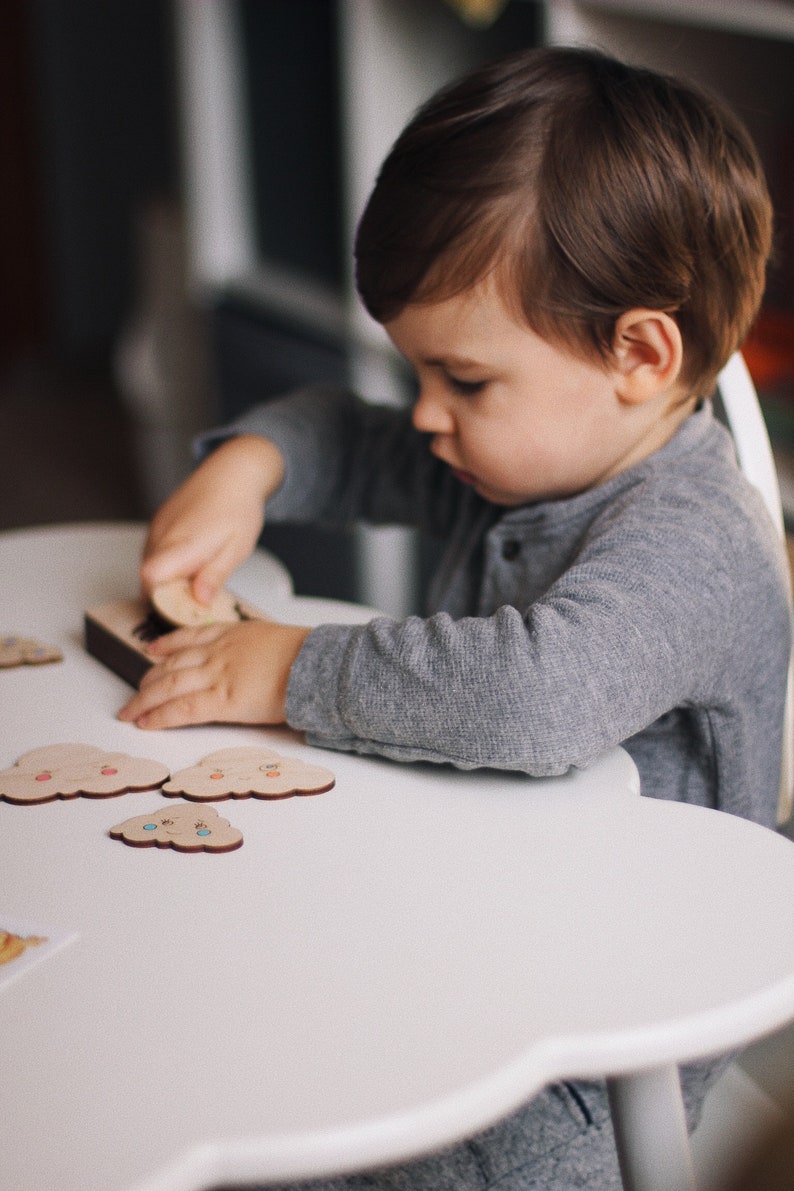 May include: A young child sits at a white, cloud-shaped table, playing with a wooden puzzle featuring colorful clouds. The puzzle pieces are arranged on the table, and the child is holding one piece in their hand.
