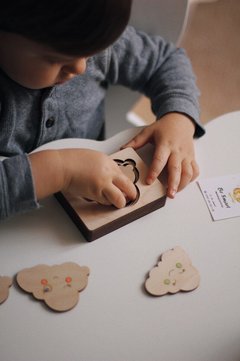 May include: A child plays with a wooden puzzle featuring a cloud shape with different facial expressions. The puzzle pieces are made of light wood and have colorful details. The child is holding a cloud piece with a smiling face.