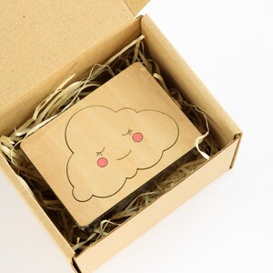 May include: A small wooden cutout of a cloud with a smiling face and closed eyes. The cloud is light brown with pink cheeks and black eyelashes. The cutout is in a brown cardboard box with shredded paper.