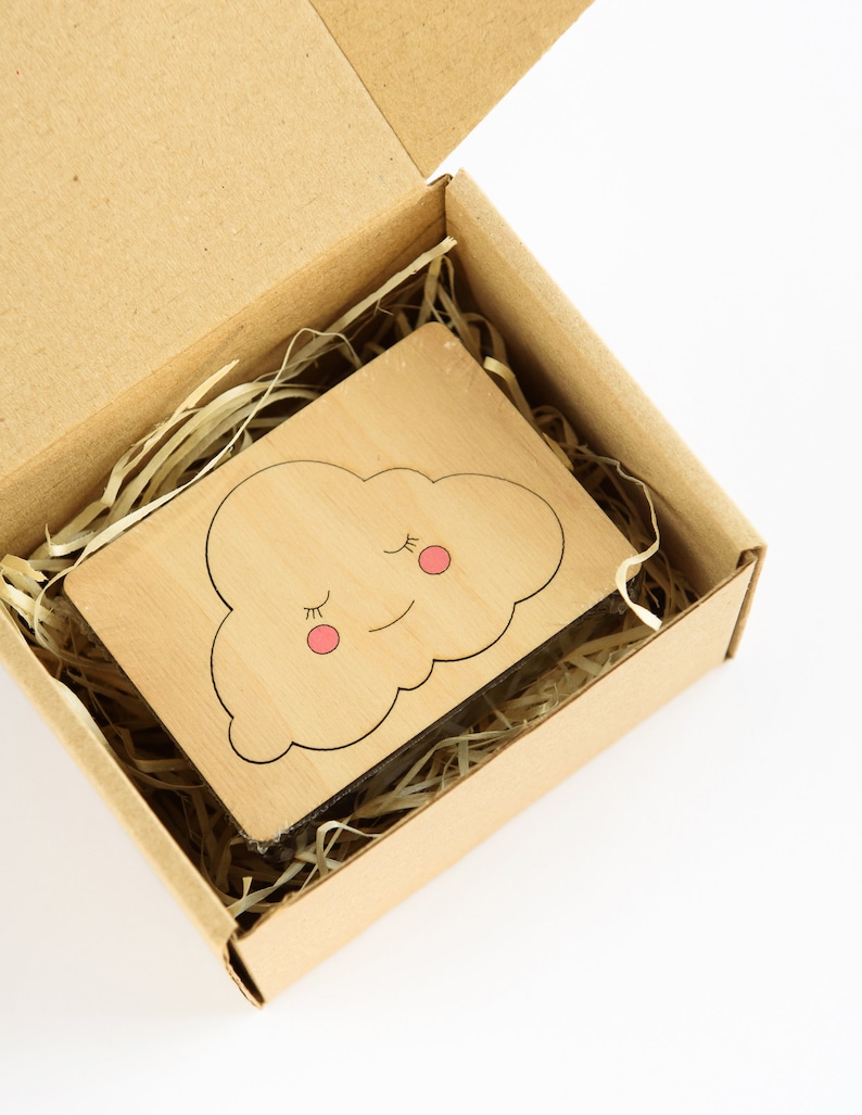 May include: A small wooden cutout of a cloud with a smiling face and closed eyes. The cloud is light brown with pink cheeks and black eyelashes. The cutout is in a brown cardboard box with shredded paper.